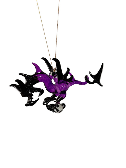 A hand-blown glass dragon ornament in purple and black, suspended by a thin gold-colored string against a white painted brick wall.